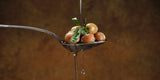 Spoon with olives and leaves being drizzled with olive oil, with drops falling against a warm brown background.